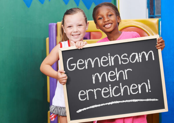 Zwei lachende Kinder halten Tafel mit dem Spruch "Gemeinsam mehr erreichen!"