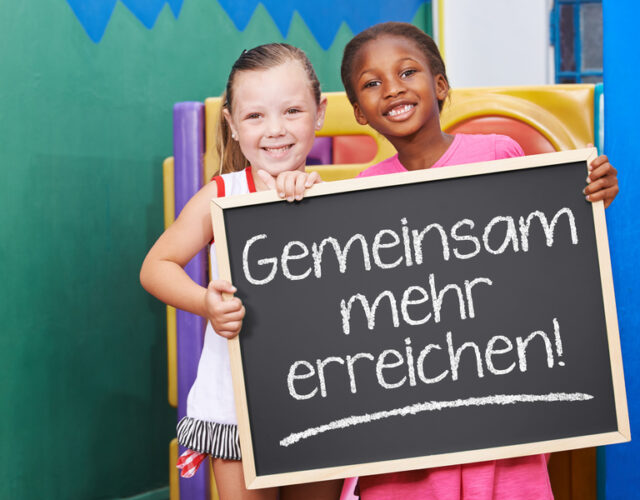 Zwei lachende Kinder halten Tafel mit dem Spruch "Gemeinsam mehr erreichen!"